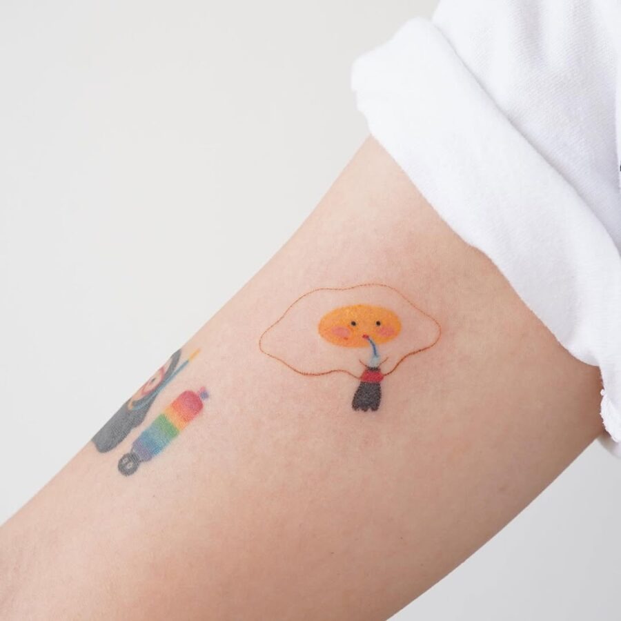 This tattoo features a playful cartoon egg character with a bright orange yolk, creating a whimsical look.