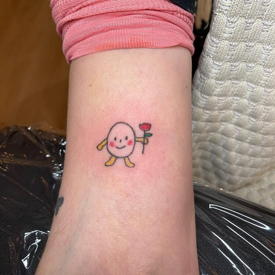 This delightful tattoo features a cheerful egg character with a whimsical smile, holding a single rose in one hand.