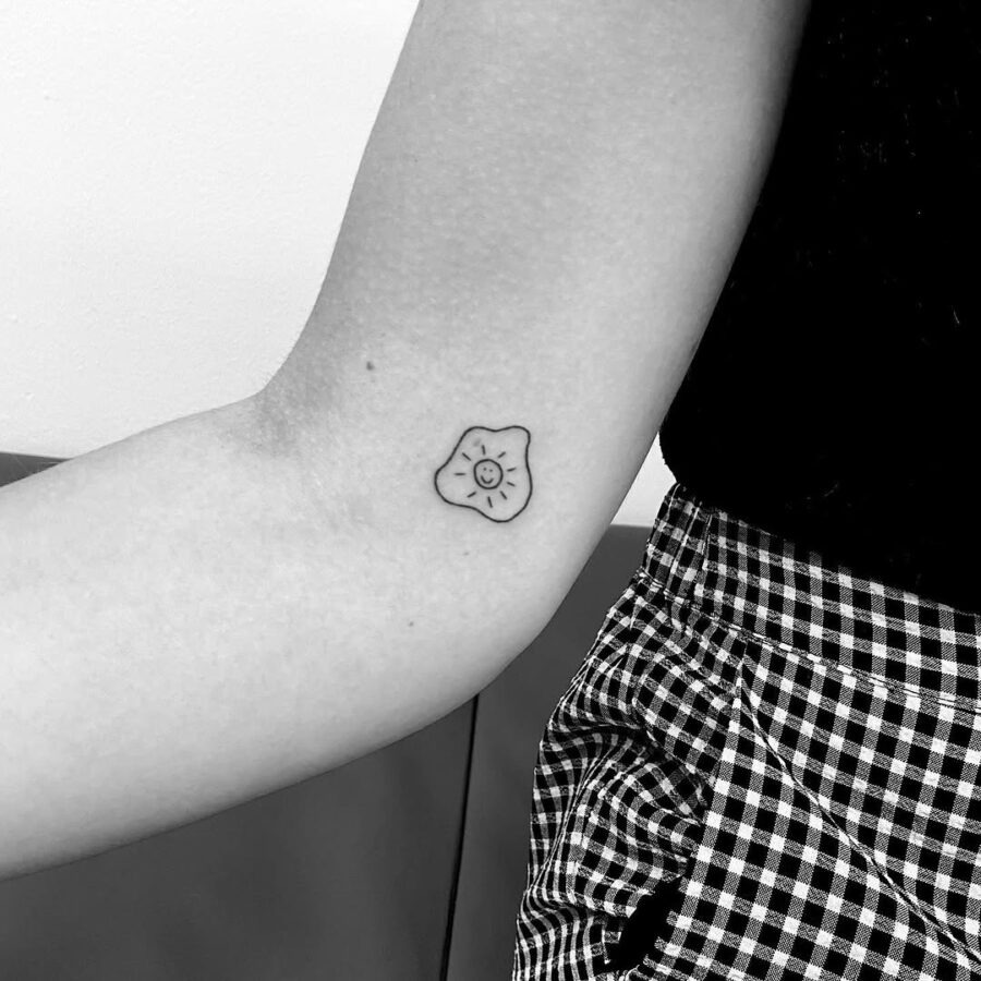 This delightful fried egg tattoo showcases a simple yet charming design featuring a smiling face.
