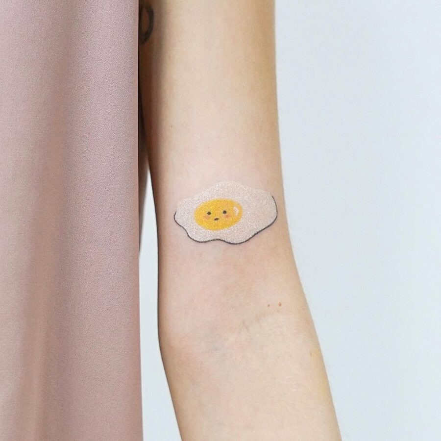 This egg tattoo showcases a simple and fun design featuring a sunny yellow yolk surrounded by a pale shell.