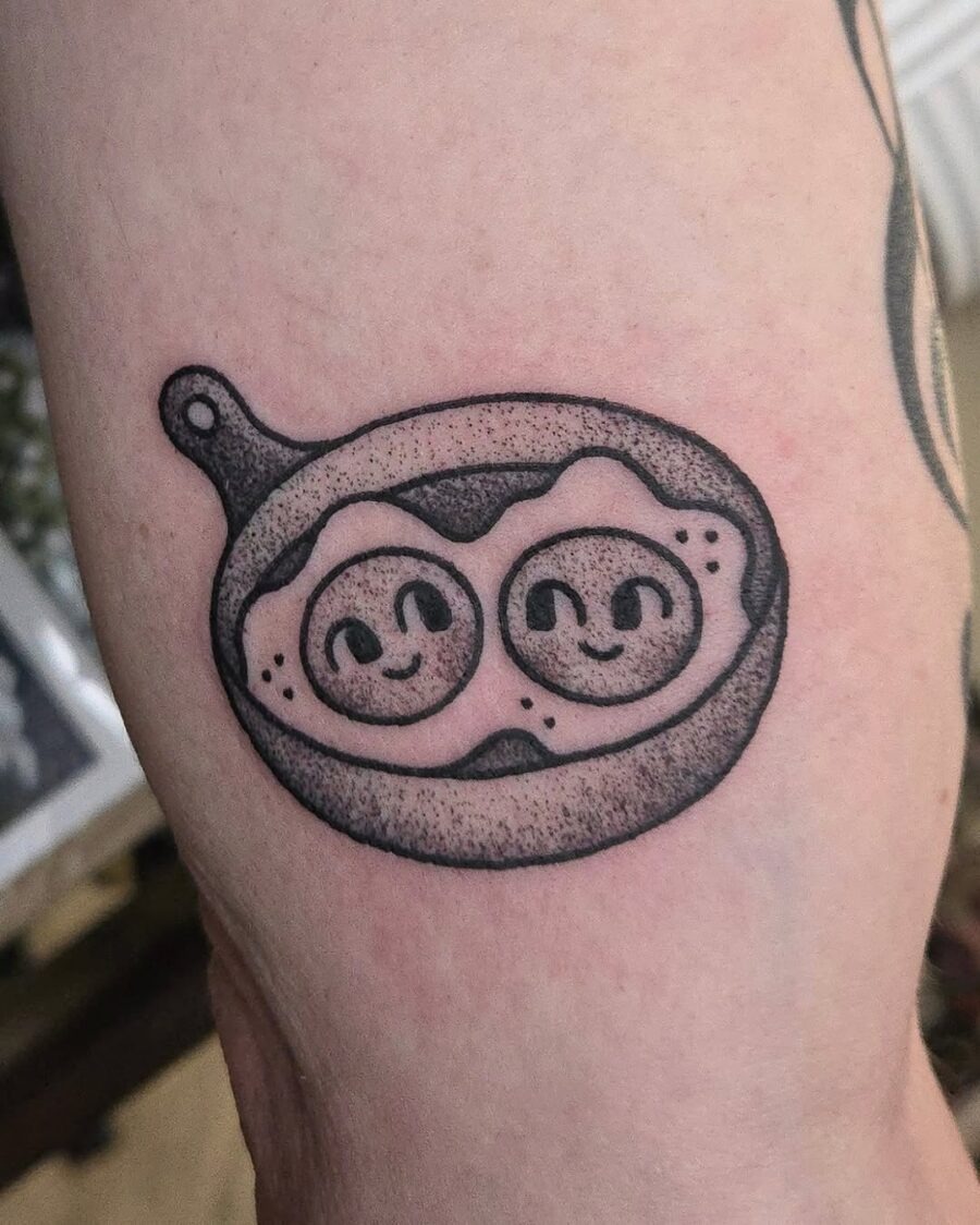 This adorable tattoo features two smiling eggs in a frying pan, exuding a playful and cheerful vibe.