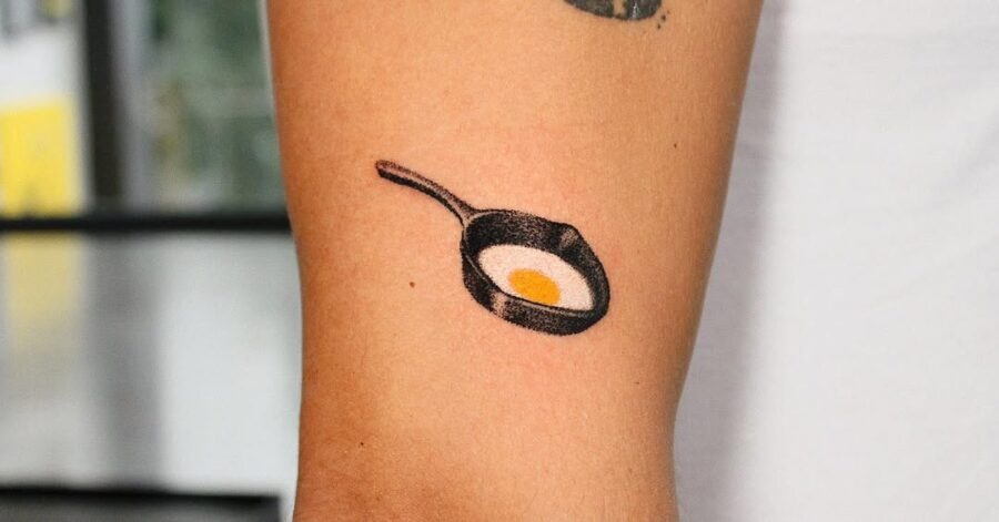 This quirky tattoo features a simple frying pan with a sunny-side-up egg, capturing a playful vibe.