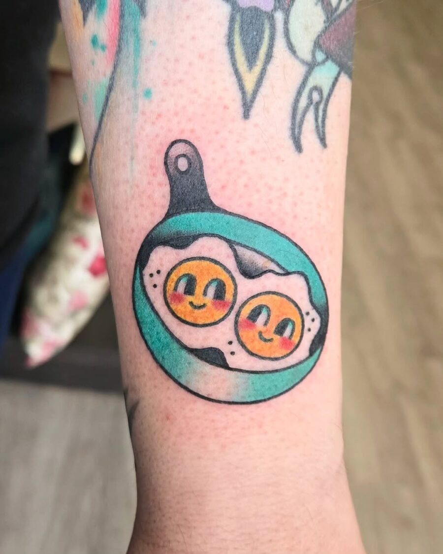 This unique tattoo features two cute, smiling eggs cooked to perfection in a skillet.