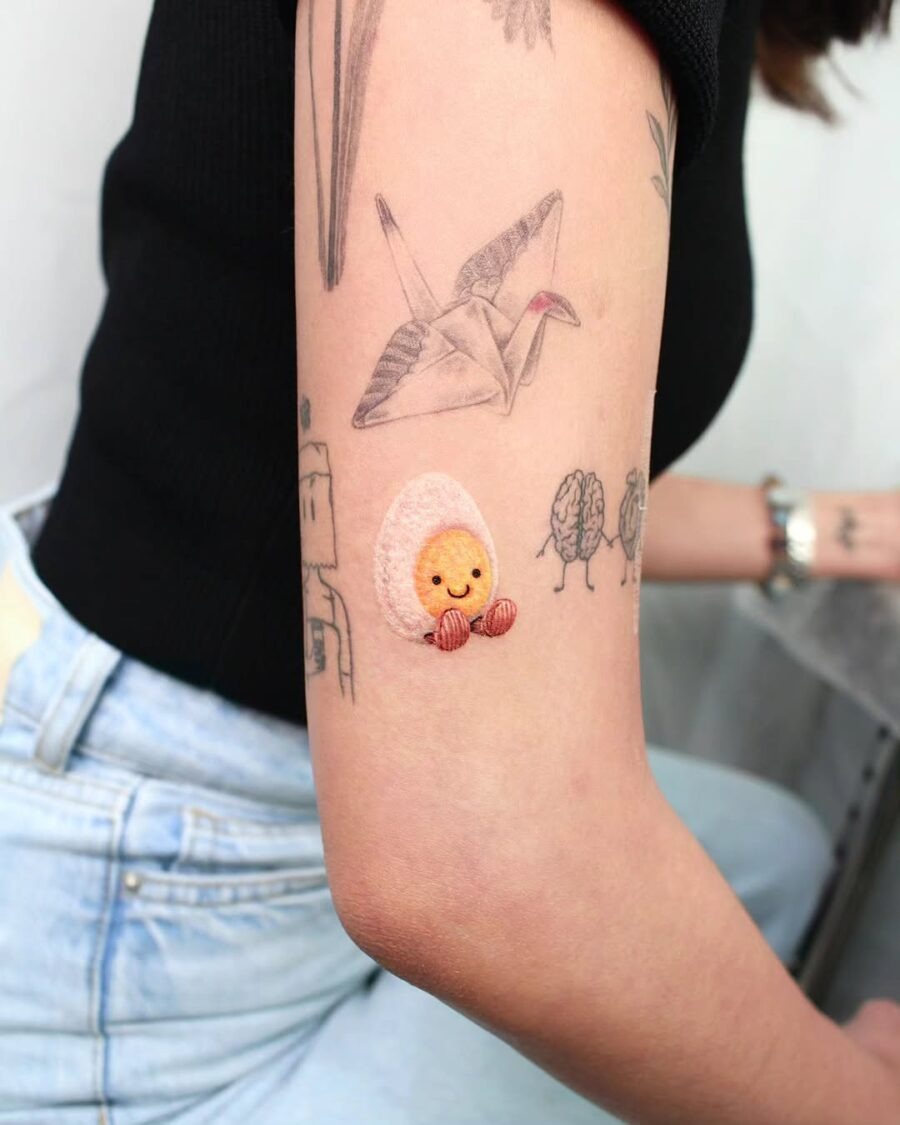 This playful tattoo features a smiling cartoon egg nestled in a pastel shell.