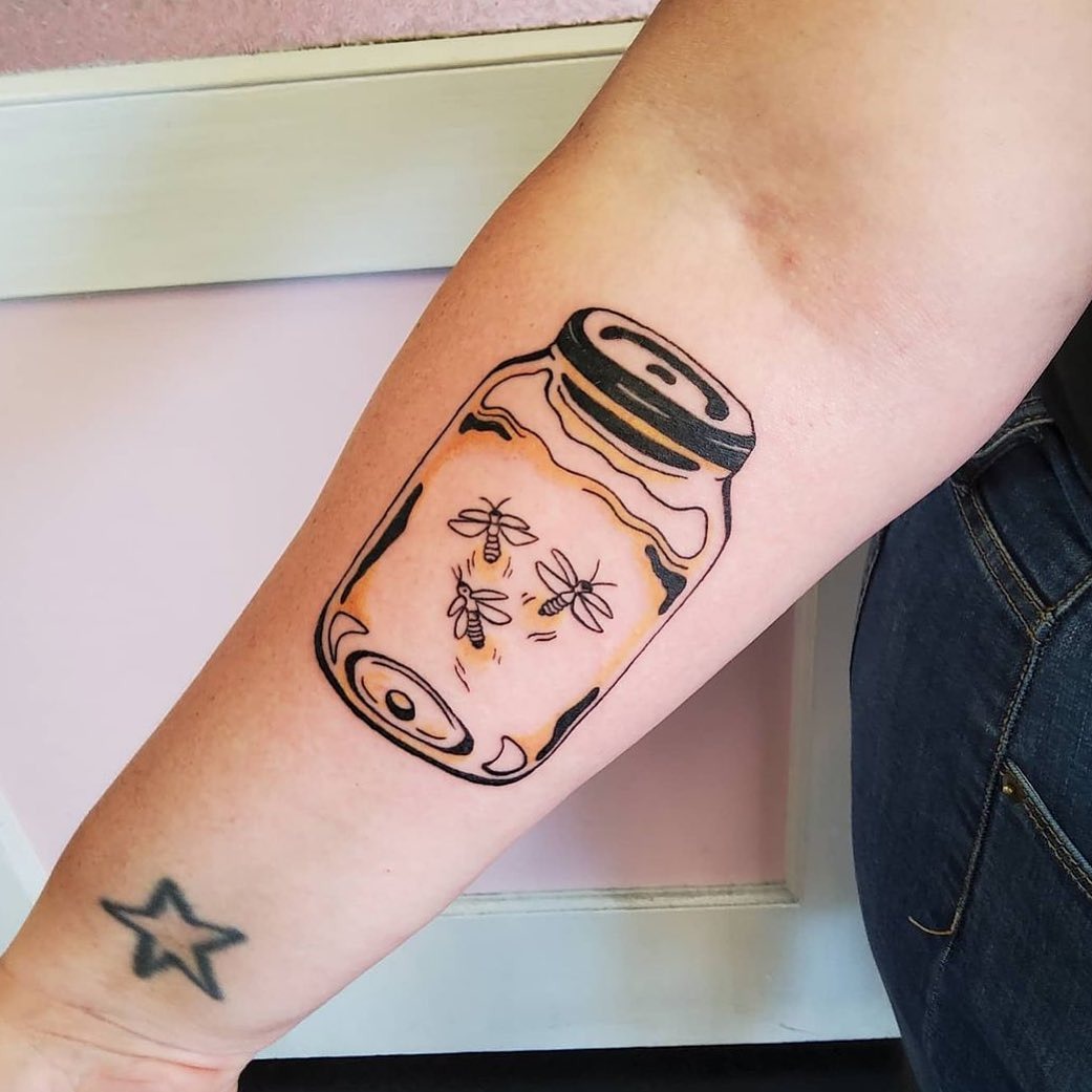 This tattoo features a whimsical jar containing fireflies, exuding a magical glow.