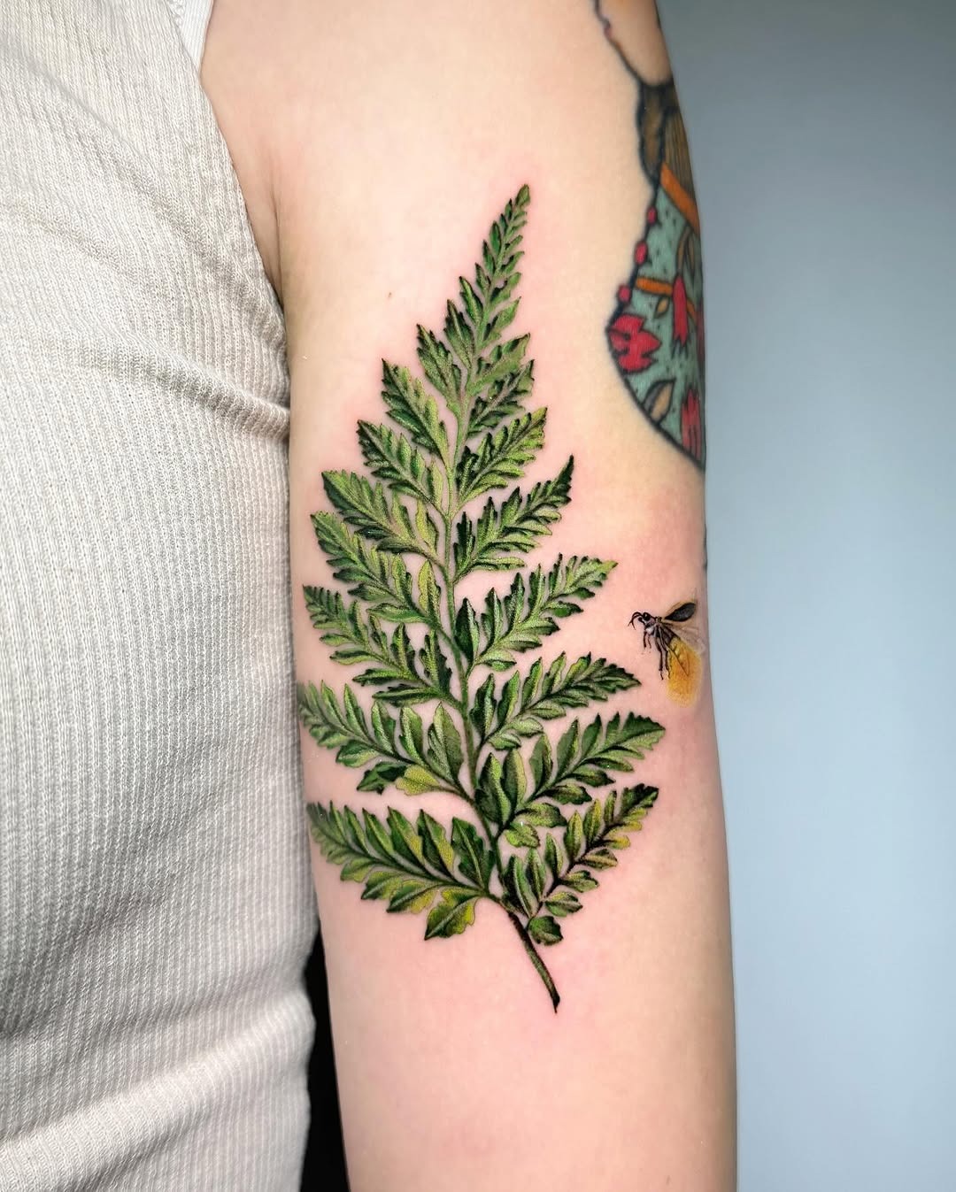 This tattoo features a green fern with a delicately detailed firefly nearby.