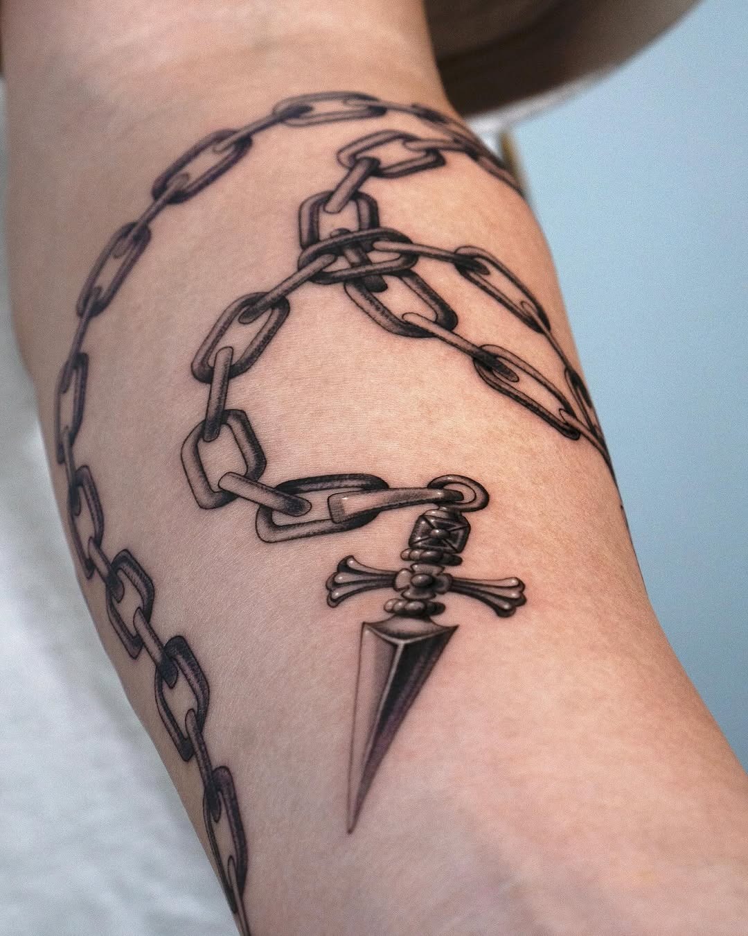 This forearm tattoo features a striking design with a chain wrapped around and a bold dagger.