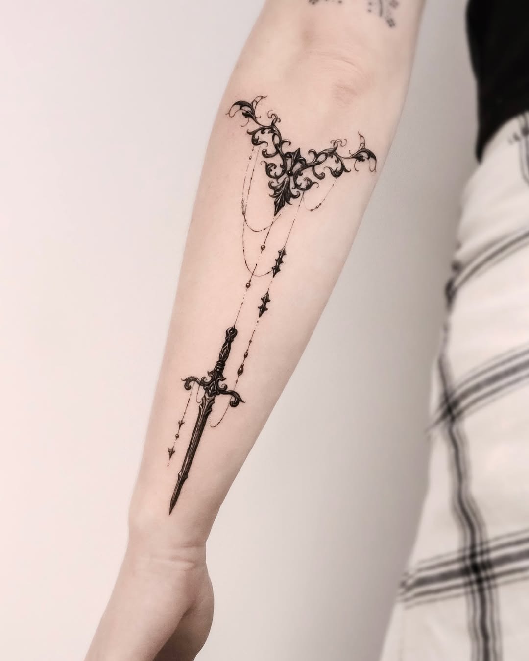 This forearm tattoo features a beautifully intricate sword design adorned with floral elements and chains.