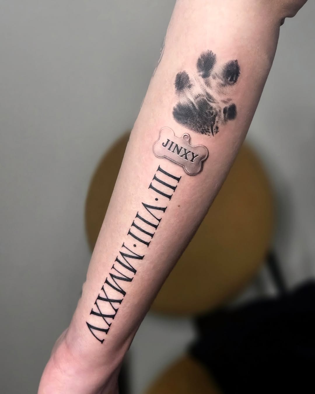 This forearm tattoo features a detailed paw print and a bone tag inscribed with the name 'Jinxy'.