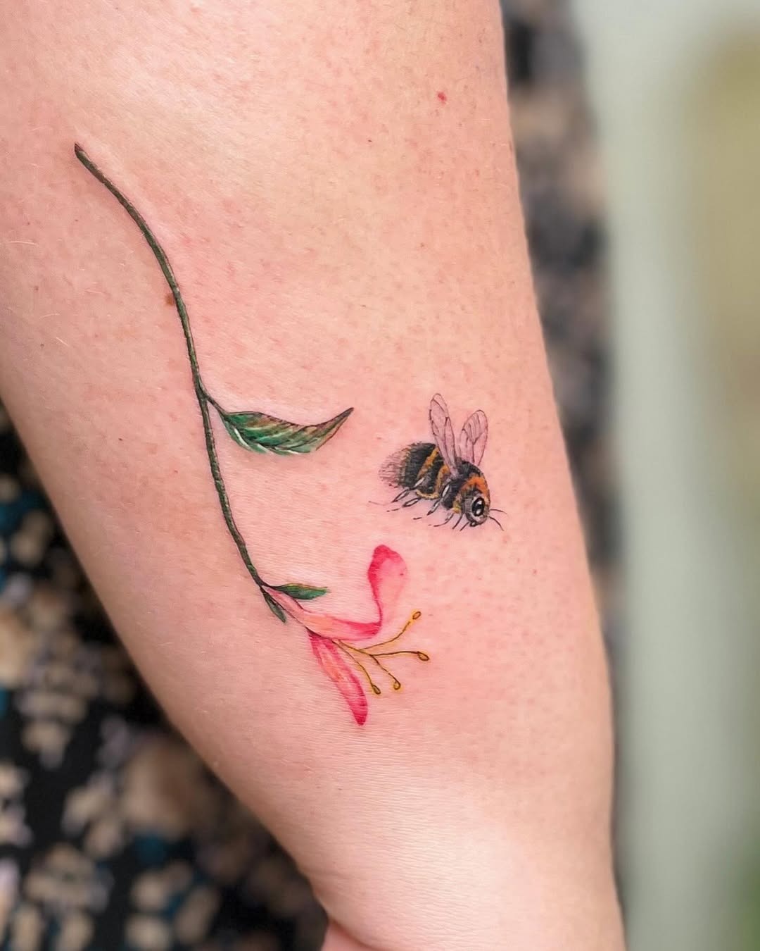 This tattoo features a delicate honeysuckle vine with pink flowers, accompanied by a detailed bee.