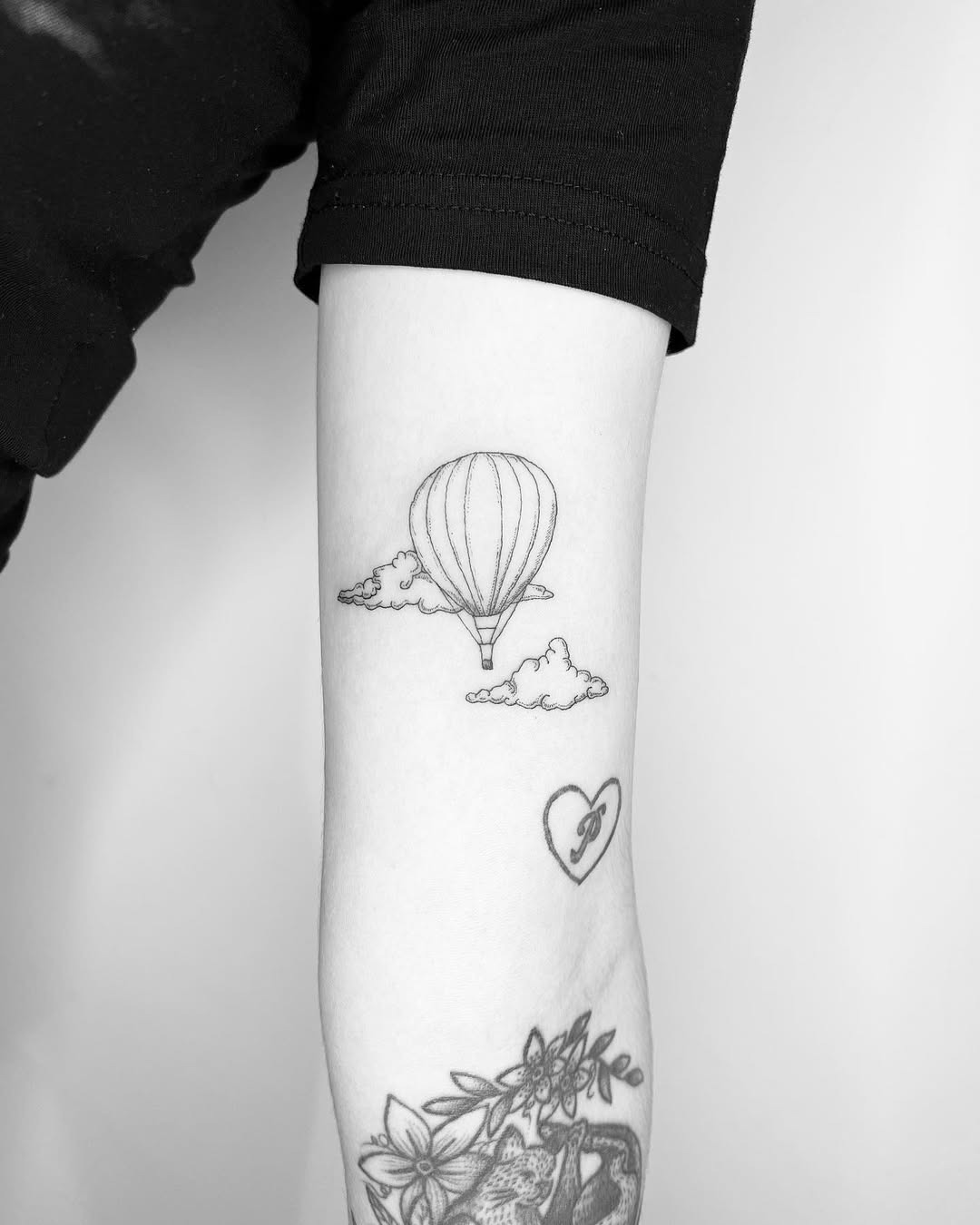 This tattoo features a simple outline of a hot air balloon soaring above fluffy clouds.