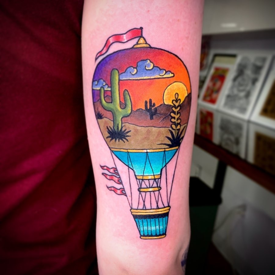 This stunning hot air balloon tattoo features a sunset backdrop, complete with playful clouds and bold cactus silhouettes.