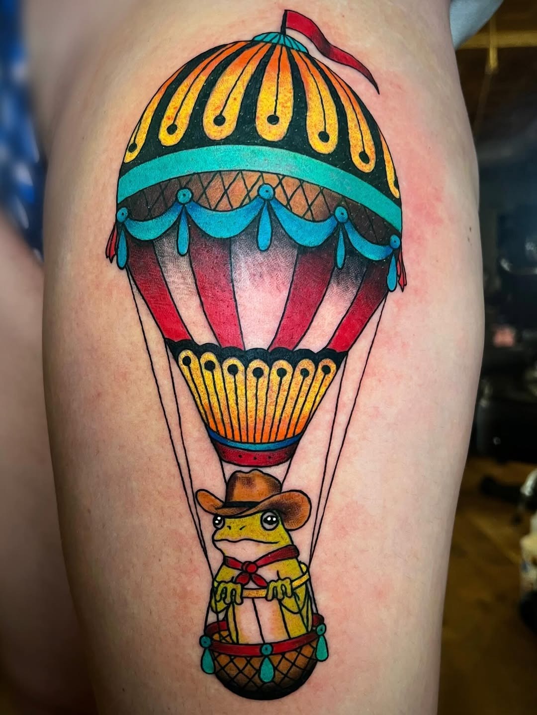 This tattoo features a cheerful frog donning a cowboy hat, nestled in a classic hot air balloon.