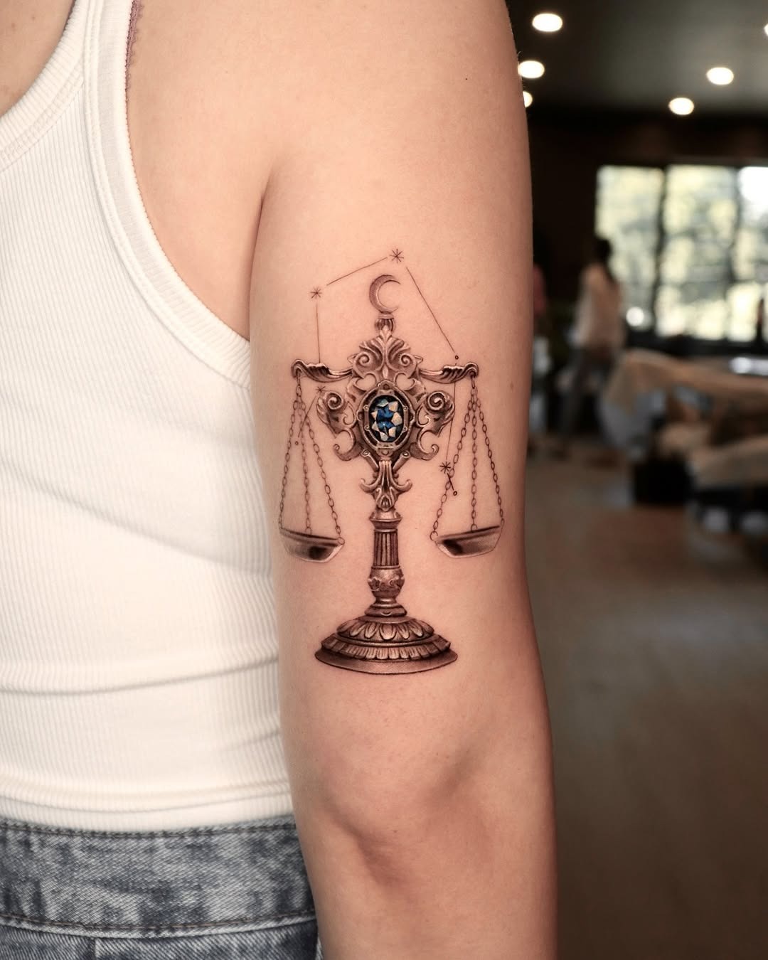 This Libra tattoo showcases a beautifully detailed scale design, highlighted by a striking blue gemstone at its center.
