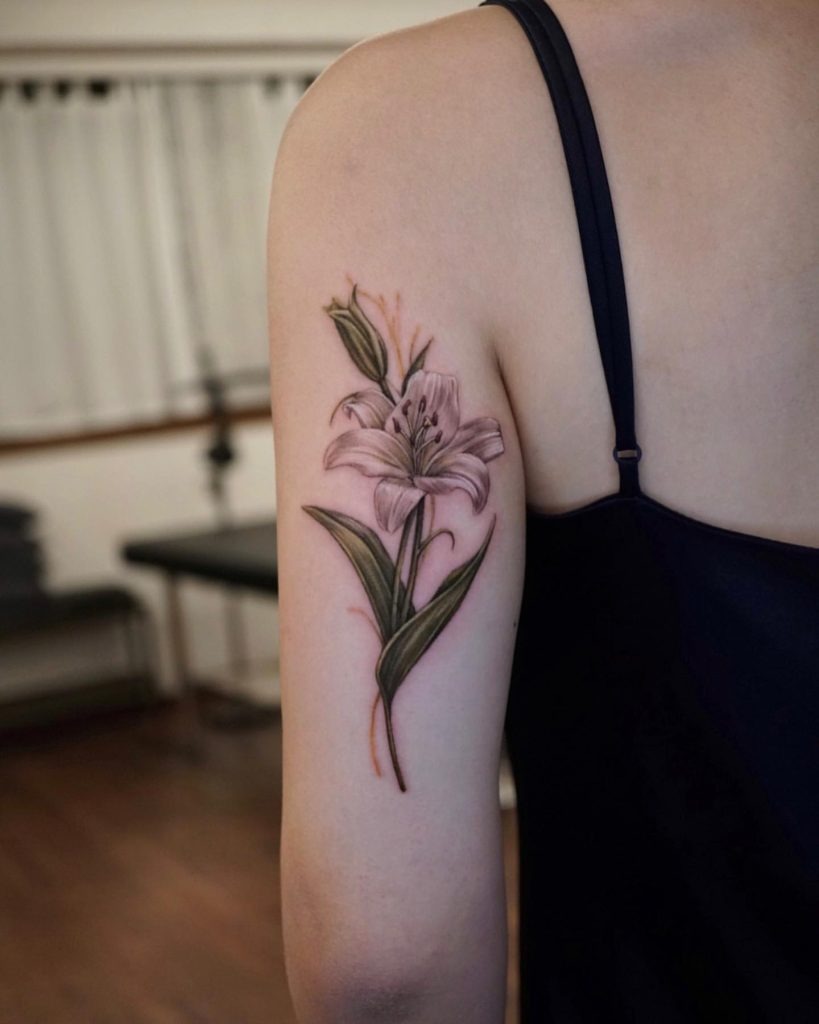 This delicate upper arm tattoo features a lily drawn with soft shading and gentle lines.