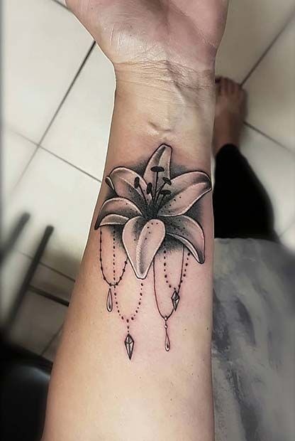 This forearm tattoo takes a unique approach by combining a large, detailed lily with beaded, hanging ornaments that drape elegantly below the bloom.