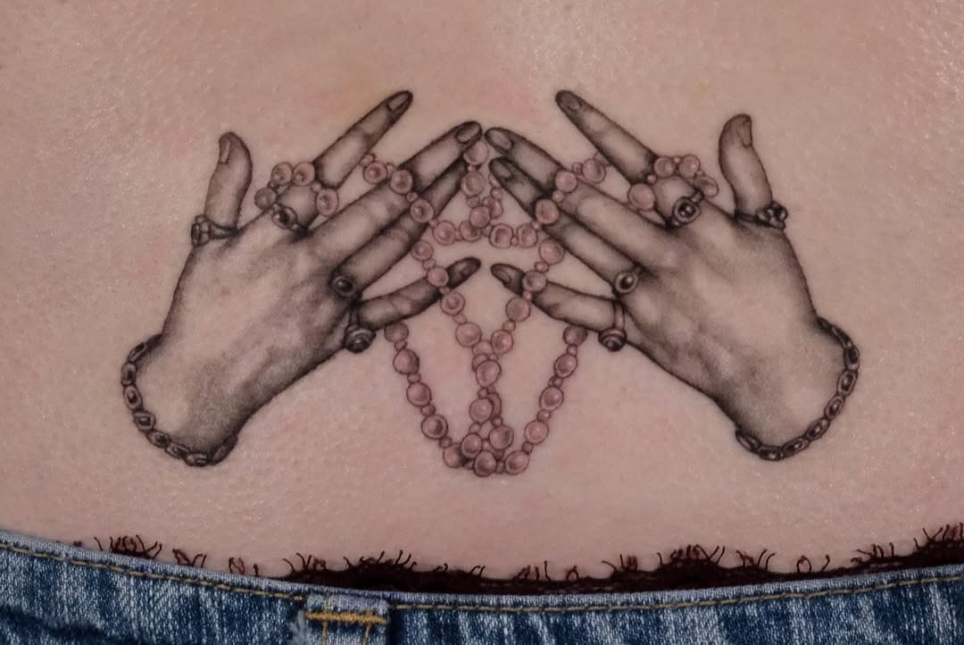 This stunning lower back tattoo features two elegantly clasped hands adorned with rings.