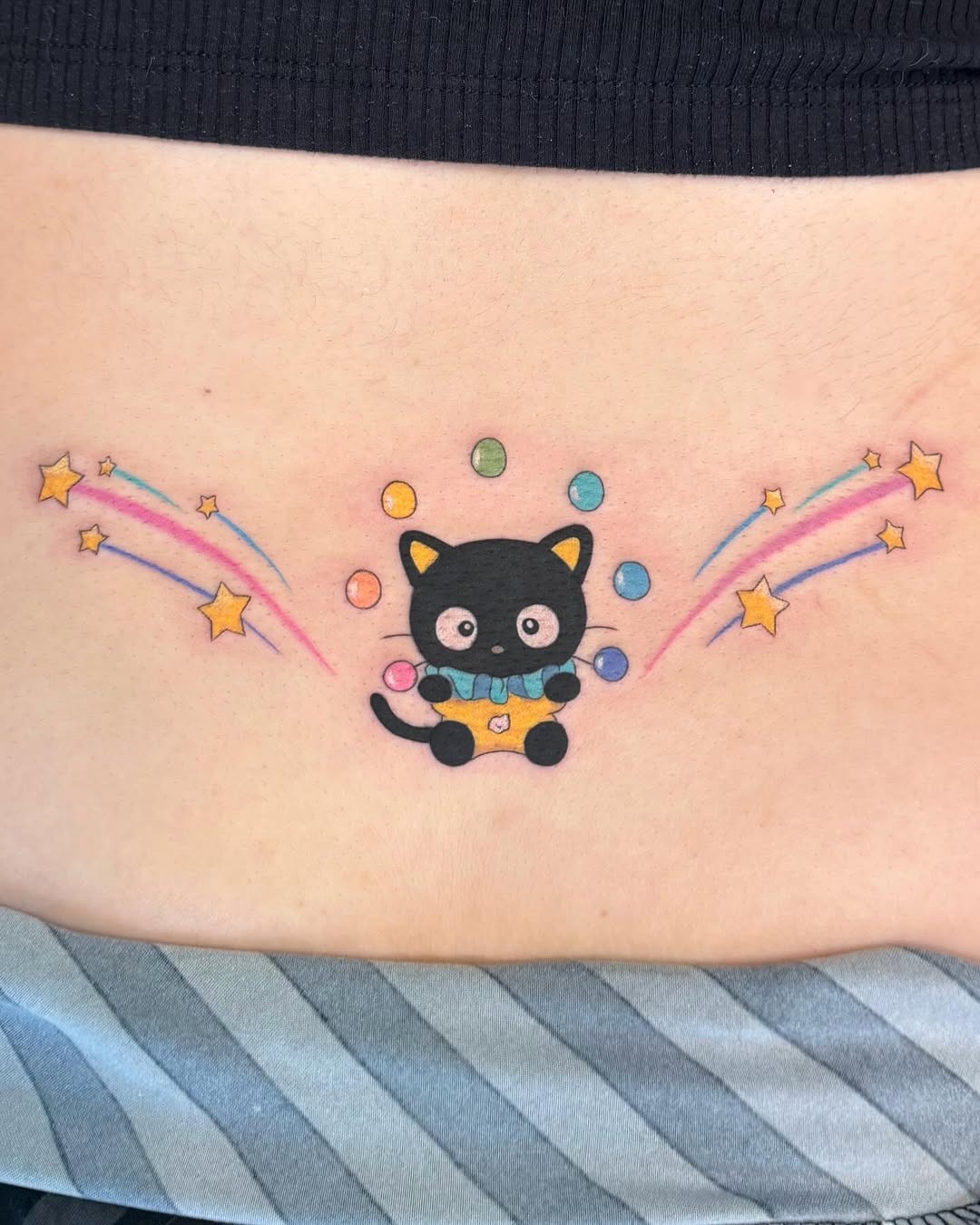 This charming lower back tattoo features a cute cat juggling colorful bubbles amidst twinkling stars.