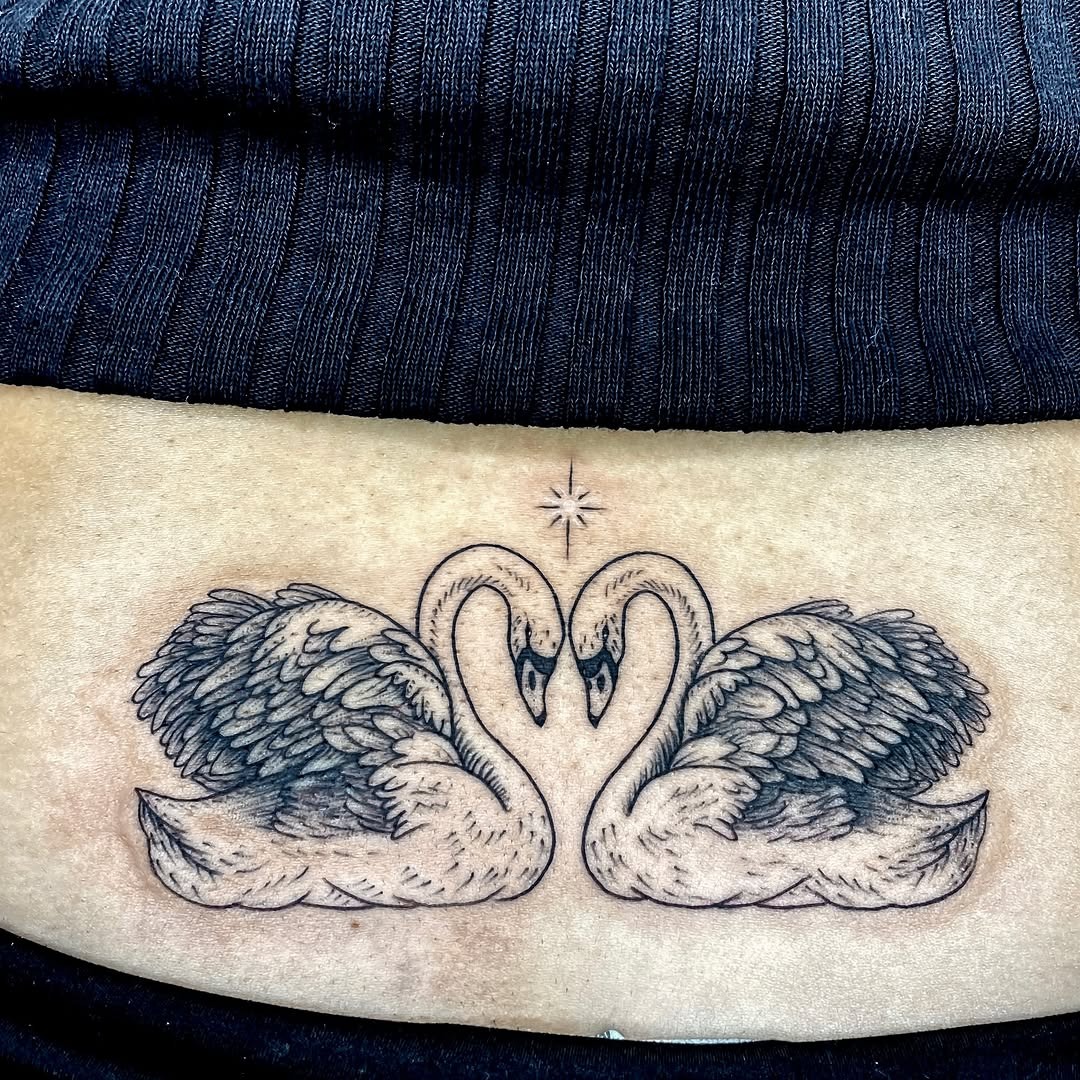 This intricate tattoo features two graceful swans with detailed wings, creating a harmonious embrace.