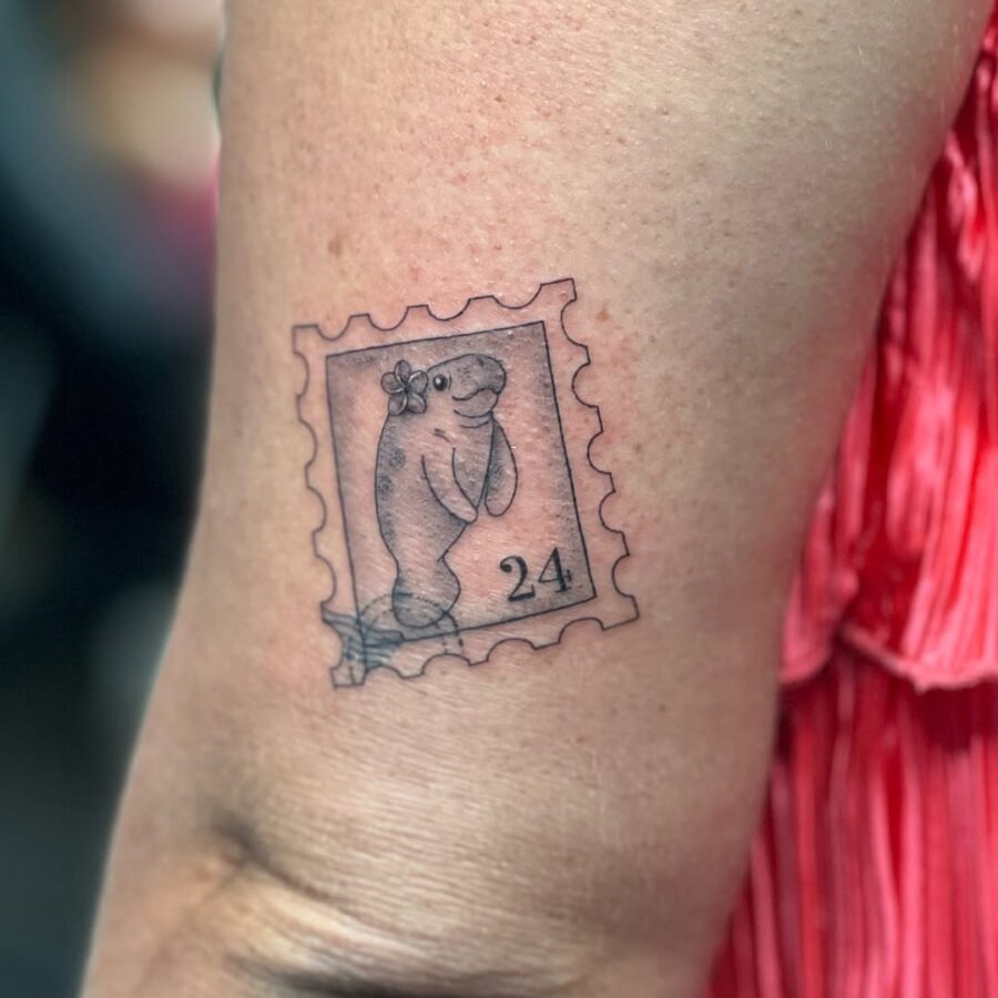 This tattoo features a delightful manatee inside a postage stamp design, giving it a whimsical, vintage vibe.
