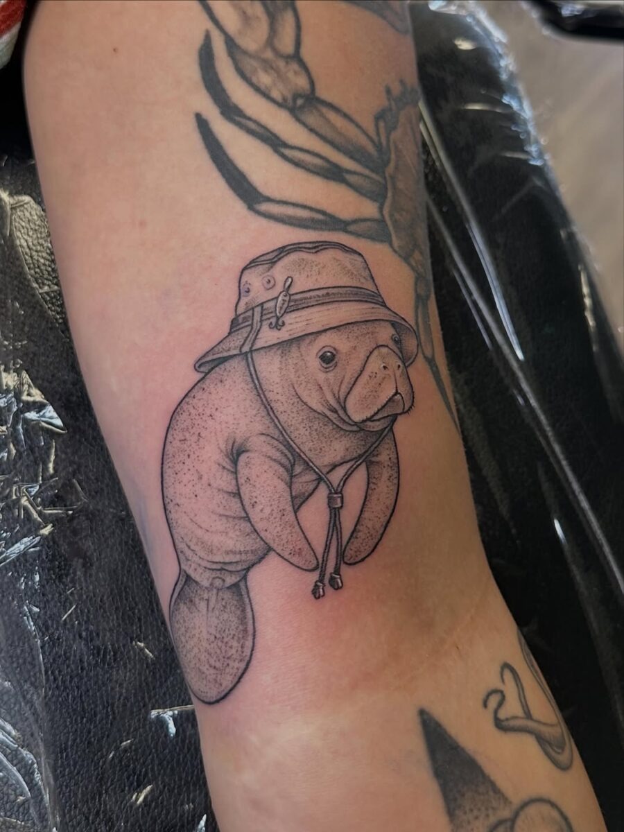 This tattoo features a charming manatee sporting a playful hat, capturing a sense of whimsy.