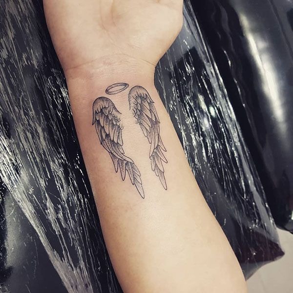 Perfect for a more dainty and minimalist approach, this wrist tattoo features petite wings delicately drawn in fine line style, topped with a small halo.