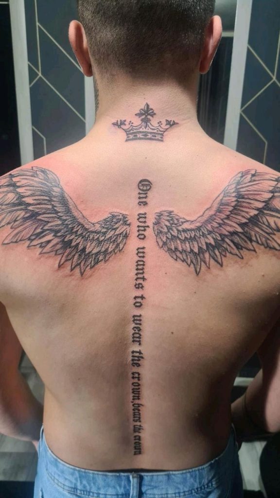A powerful angel wings tattoo on back that features a crown at the neck and a vertical quote down the spine: "One who wants to wear the crown bears the crown." This is a visual metaphor for responsibility and power, blended with regal and celestial motifs.