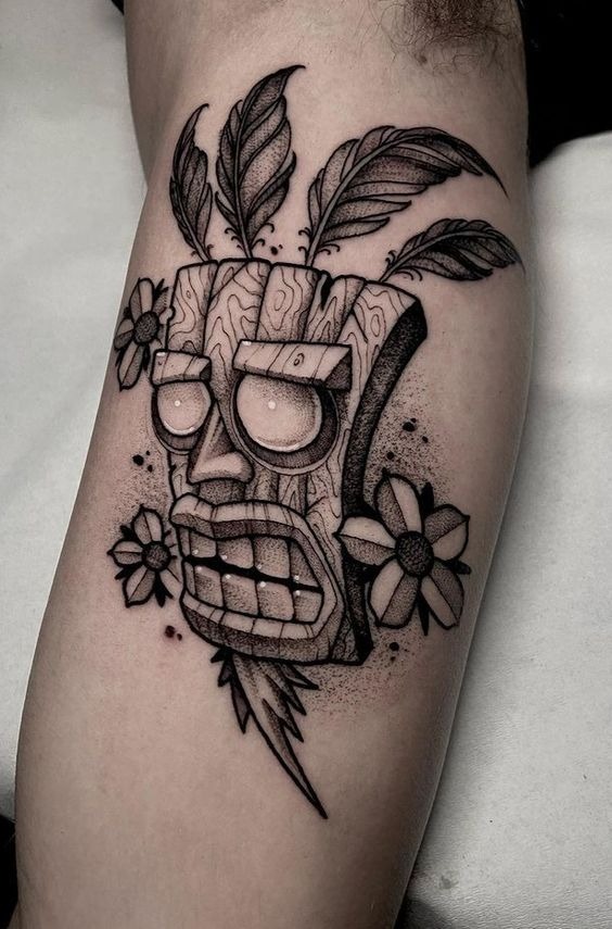 This tattoo explodes with character, a tiki-style wooden mask with wide cartoonish eyes, feather plumes, and daisy details.