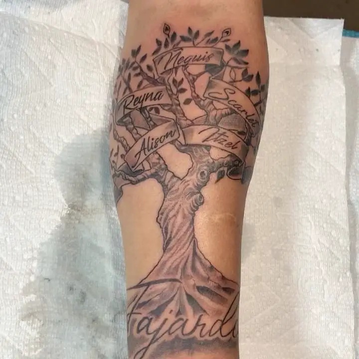 Spanning the forearm, this tattoo features a detailed tree with carefully scripted names such as Reyna, Nequis, and Alison on banners wrapping around the branches.