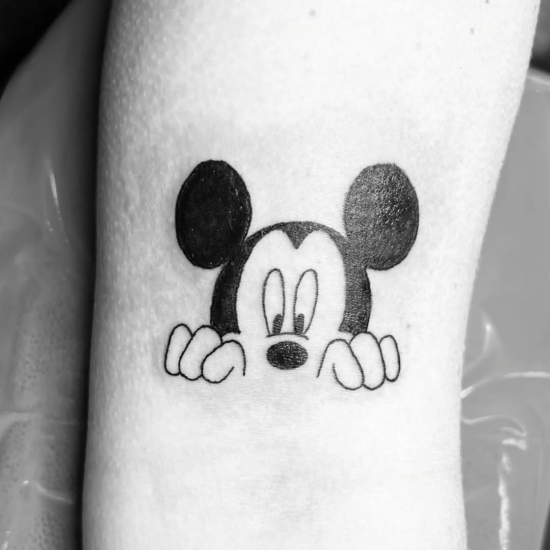 This tattoo features a playful depiction of Mickey Mouse peeking over the skin, showing his iconic round ears and cheerful expression.