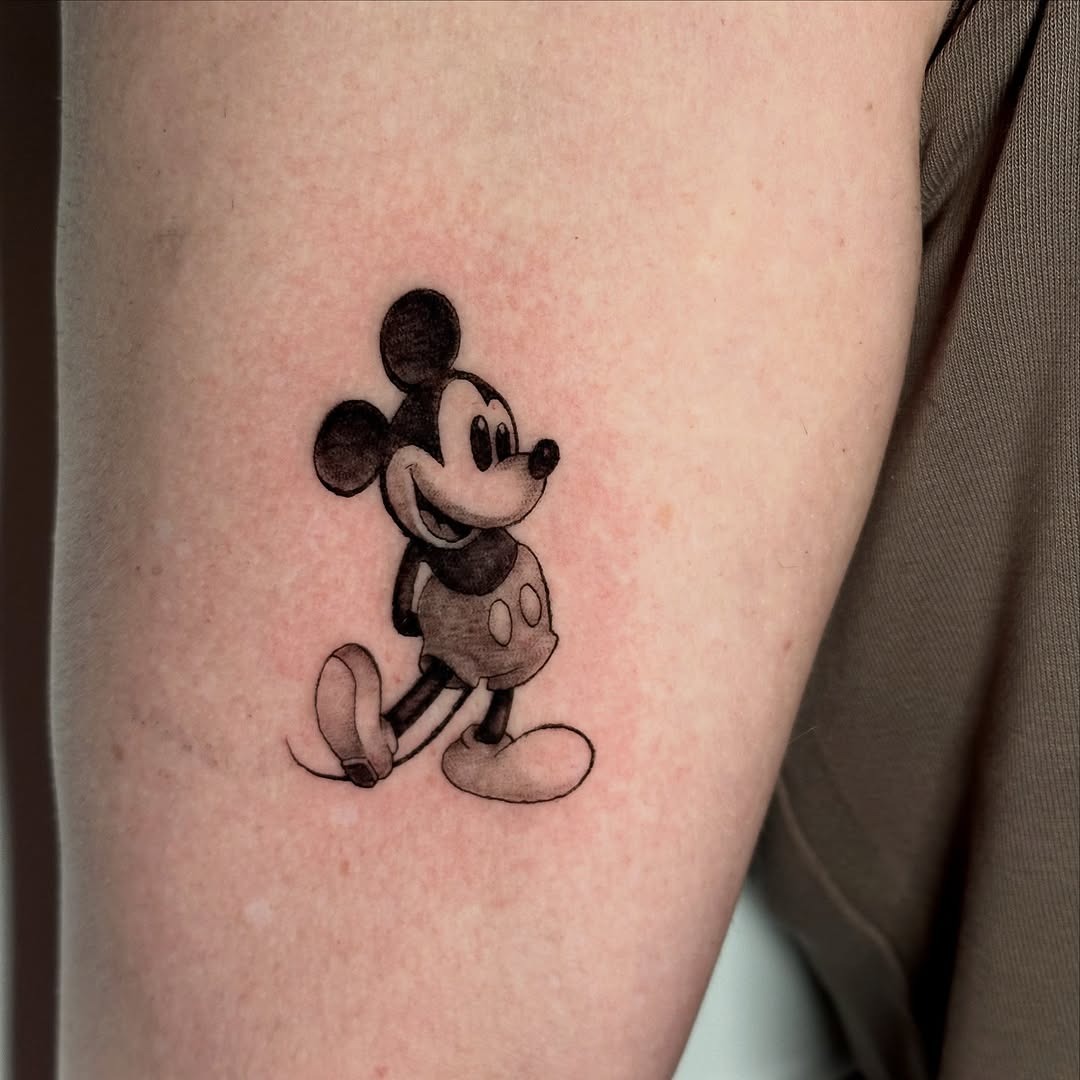 This Mickey Mouse tattoo features the iconic character in a playful pose, rendered beautifully in black and grey ink.