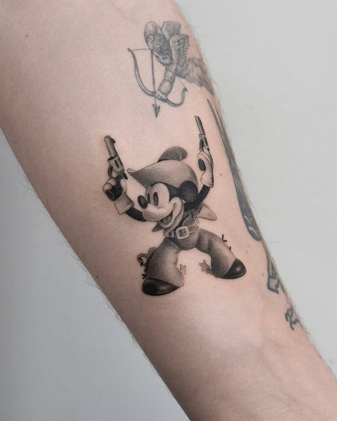 This tattoo features a playful rendition of Mickey Mouse dressed as a cowboy, brandishing two six-shooters.