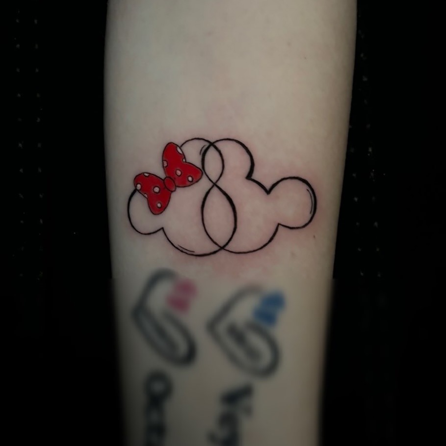 This tattoo features the iconic Mickey Mouse silhouette intertwined with an infinity symbol, accentuated by a classic red bow.