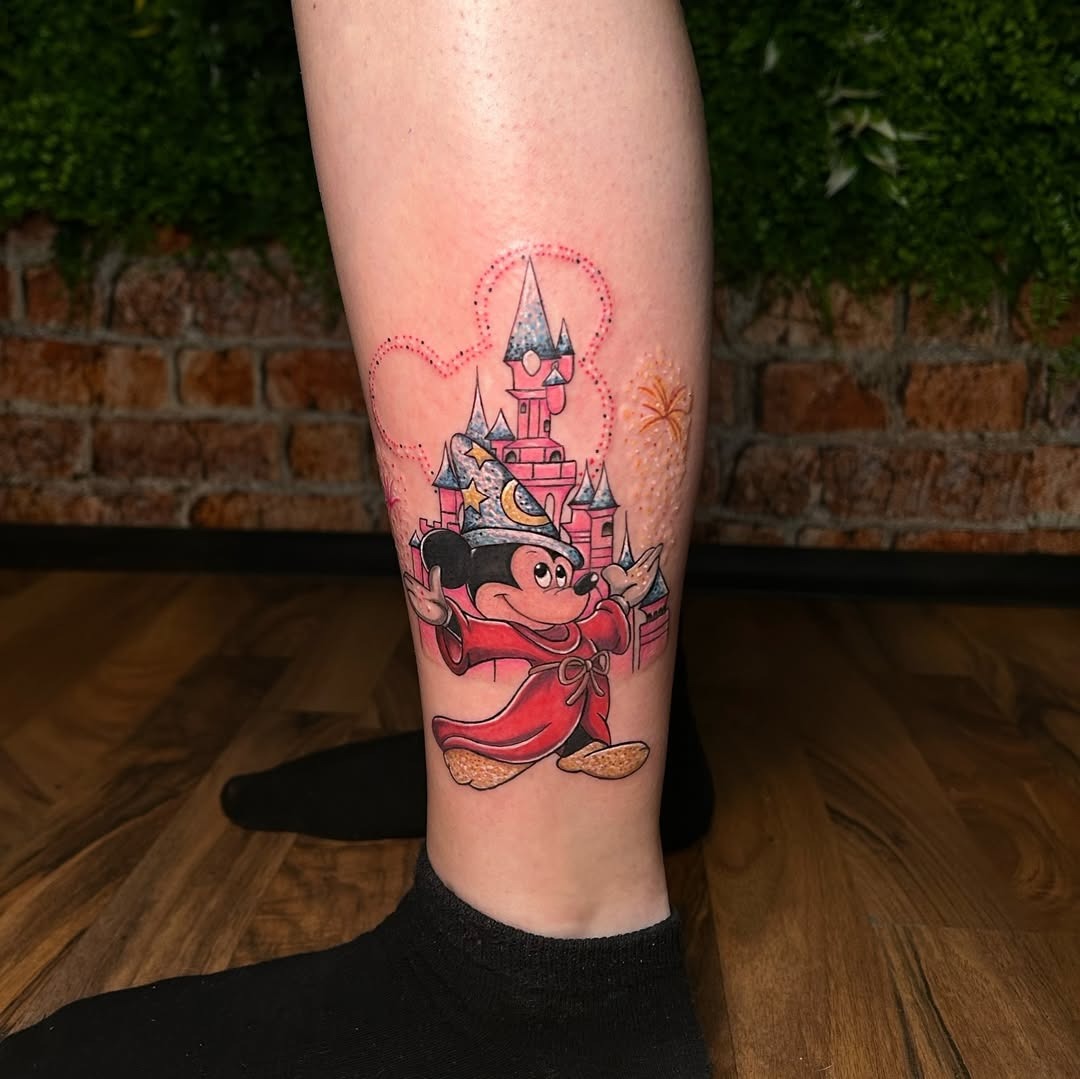 This colorful tattoo features Mickey Mouse in a playful pose, adorned with a crown, in front of a splendid castle.