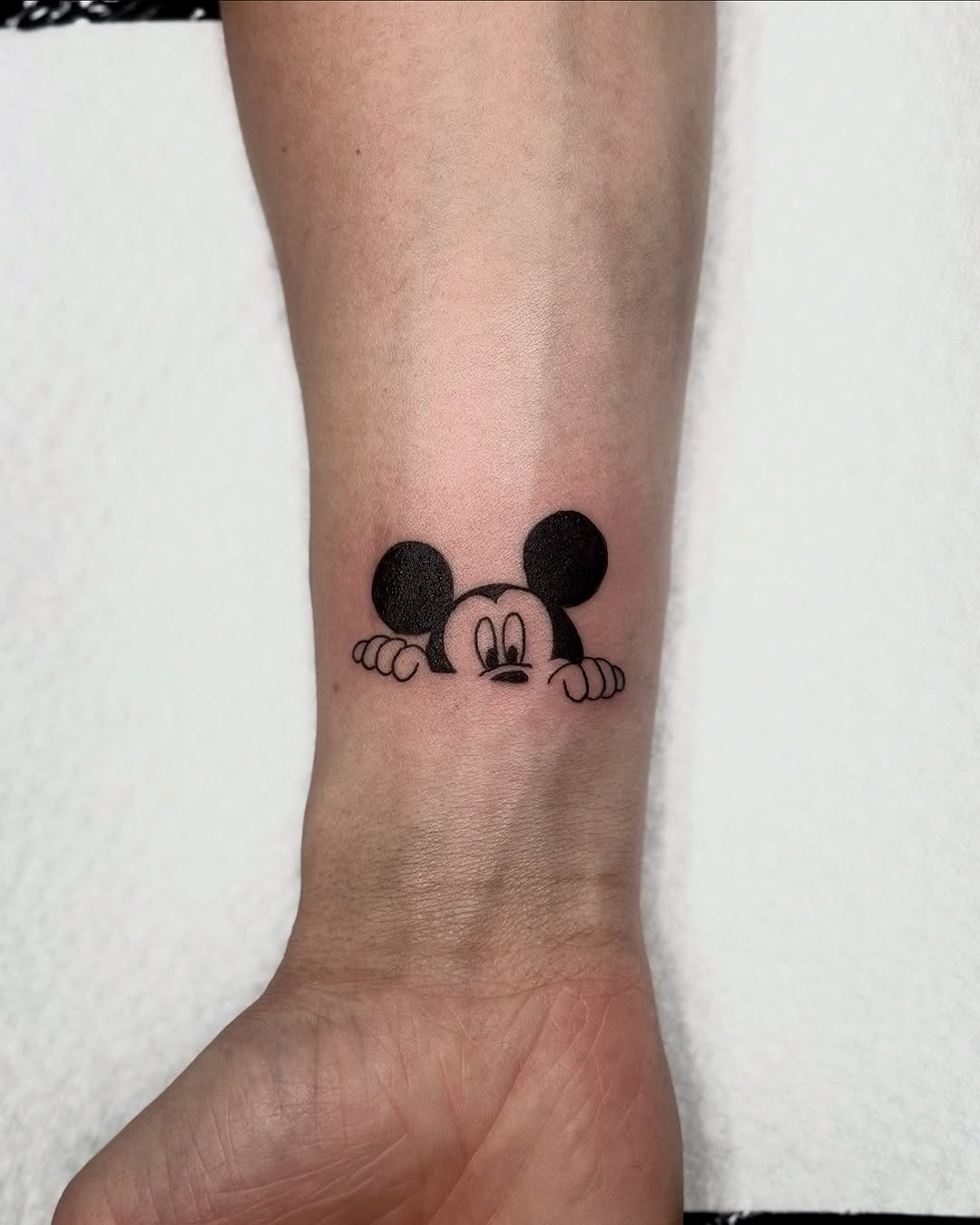 This whimsical tattoo features Mickey Mouse peeking playfully from the underside of the wrist.