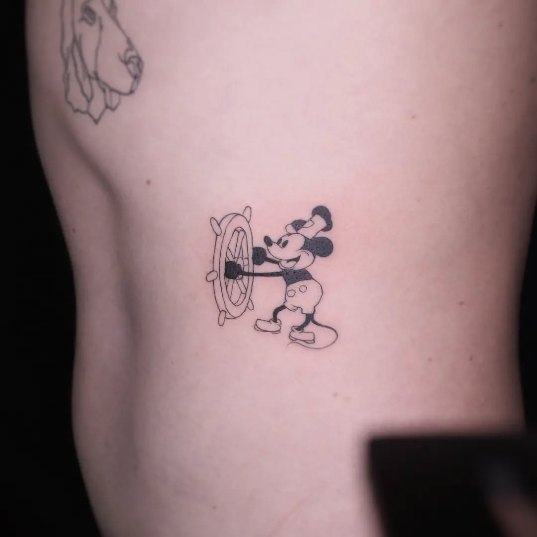 This tattoo showcases Mickey Mouse playfully grasping a steering wheel, embodying a sense of adventure and nostalgia.