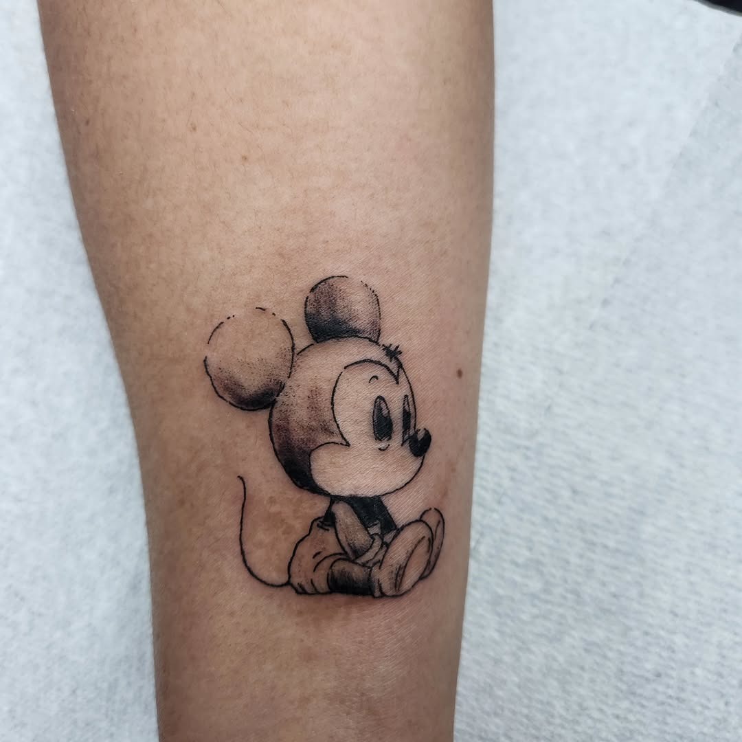 This charming tattoo features a classic portrayal of Mickey Mouse, sitting playfully in a relaxed pose.