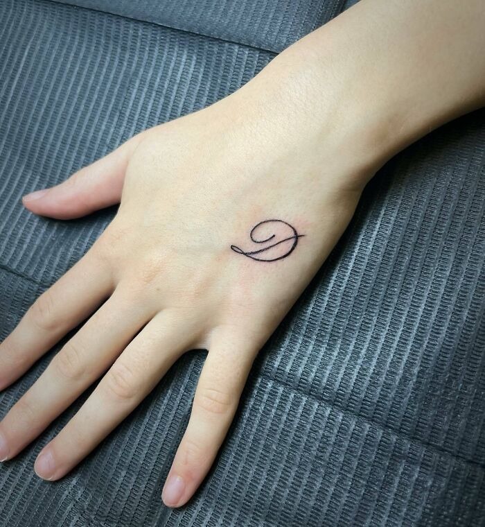 This tattoo showcases a graceful swirl transforming into a letter G, etched delicately on the back of the hand.