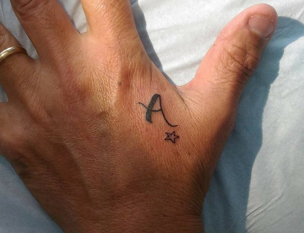 This tattoo features a bold letter A accompanied by a small star on the hand, close to the thumb.