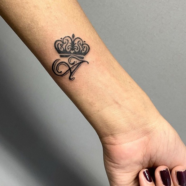 A more ornate design, this tattoo combines a cursive letter A with an intricately detailed crown placed above it on the wrist.