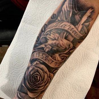 Featuring a dove in flight above a detailed rose, this tattoo incorporates a ribbon with the name "Maria" and significant dates.