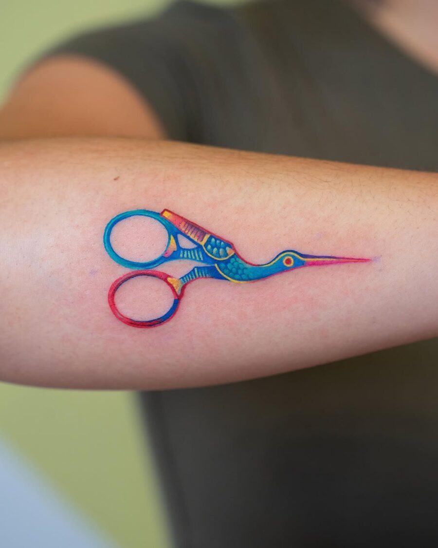 This vivid tattoo features a pair of scissors cleverly transformed into a colorful bird.