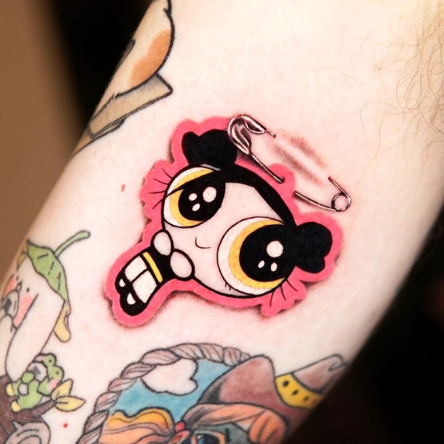 This tattoo features a playful rendition of a Powerpuff Girl, marked by bright colors and a quirky safety pin.