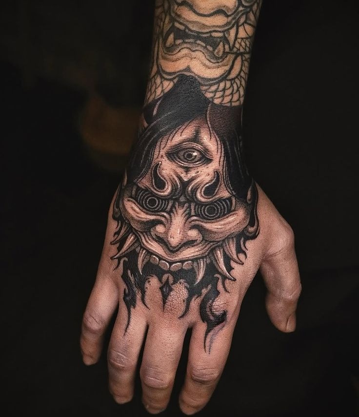 This hand tattoo features an otherworldly oni mask with a third eye, etched with swirling patterns and intense expression.
