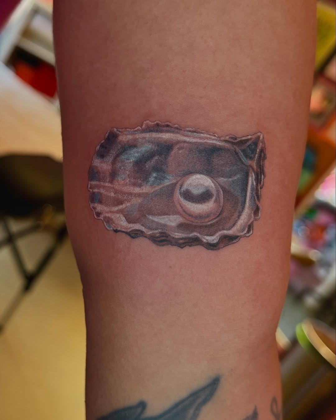 This tattoo features a beautifully rendered oyster with a gleaming pearl inside, showing intricate details.