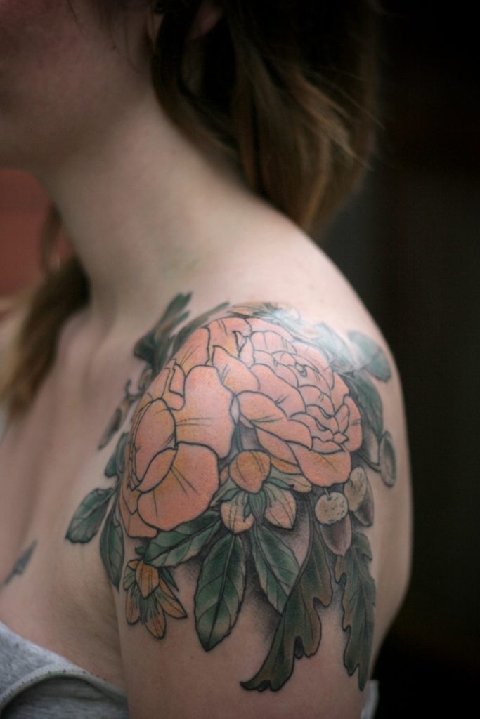 This shoulder cap tattoo features a large, bold flower with bright orange petals and green leaves, creating a striking women flower design.