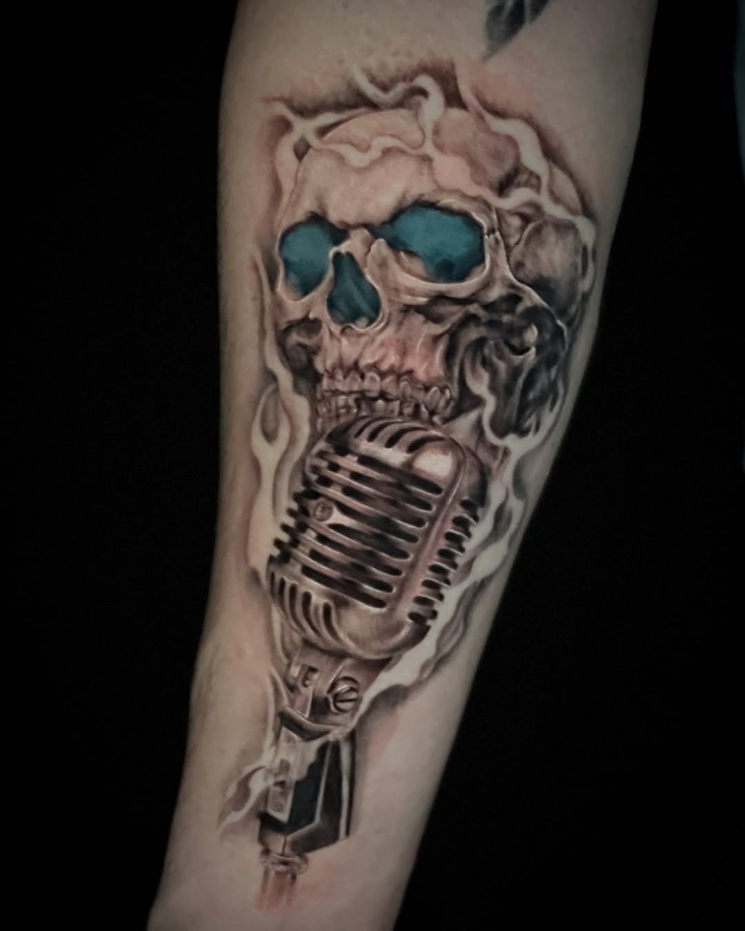 This tattoo features a skull with turquoise eyes, intertwined with a vintage microphone.