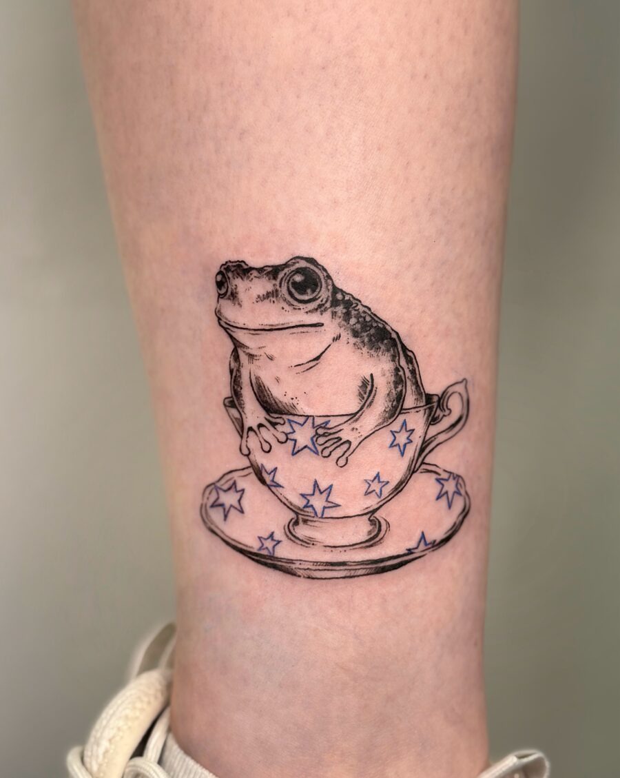 A playful frog nestled in a teacup Tattoo Design