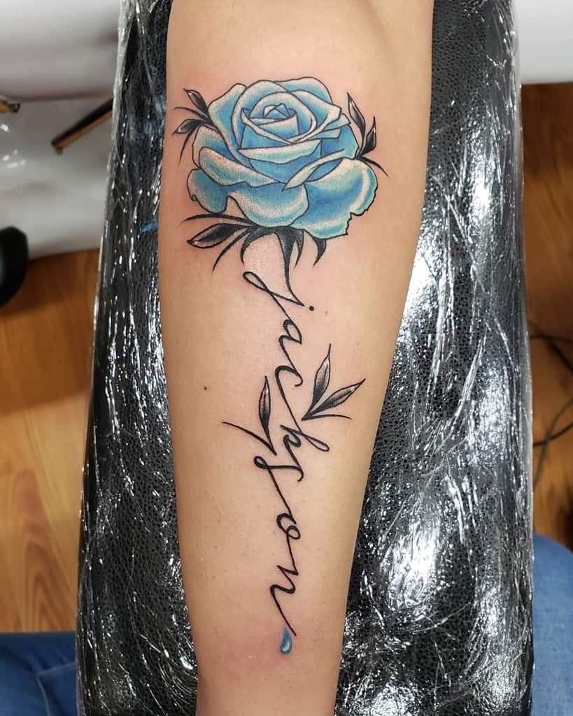 This forearm tattoo showcases a delicate blue rose paired with elegant script spelling out "Jackson" and a small blue drop, adding a touch of sentimental value.