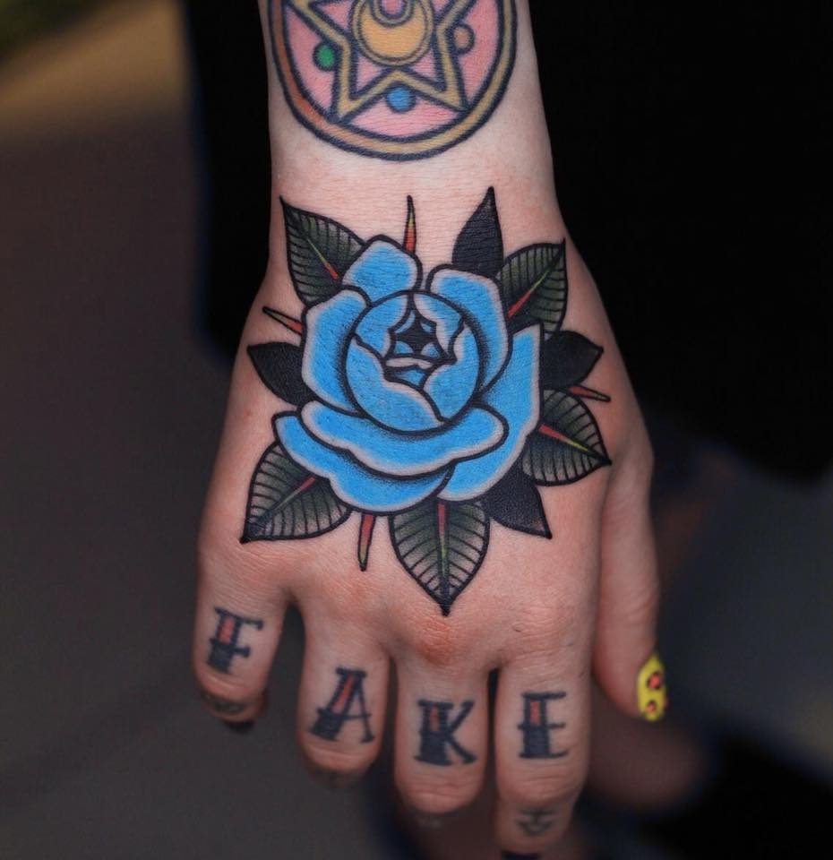 This traditional hand tattoo stands out with its bold outlines and a radiant blue rose at its center, surrounded by vividly colored leaves.